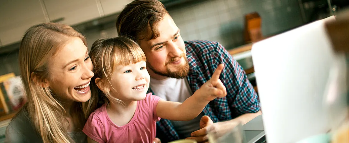 Junge Familie mit Kind lächelt gemeinsam am Laptop am Frühstückstisch