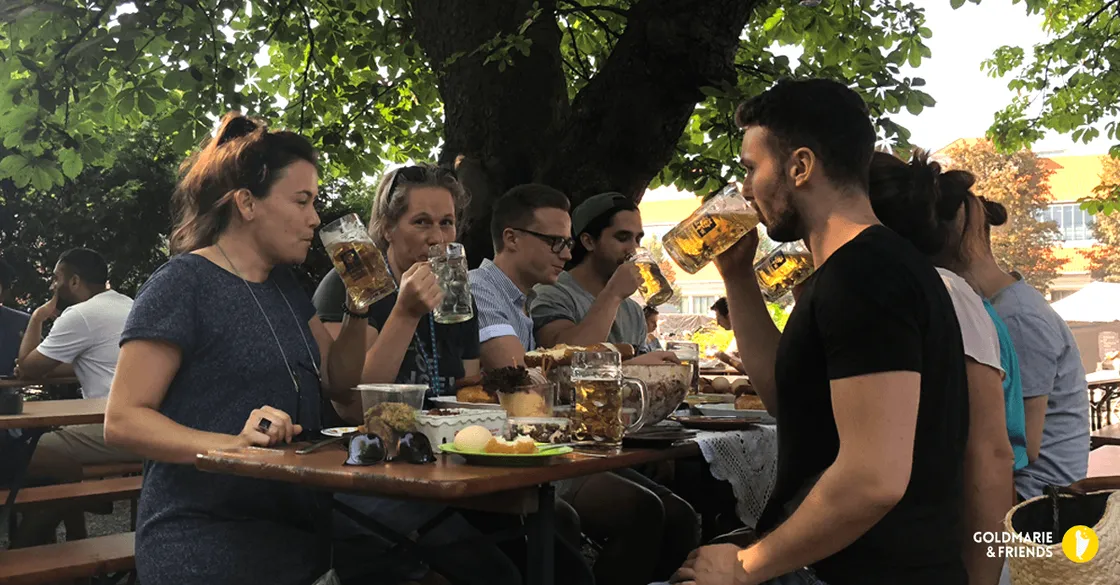 Menschen sitzen im Biergarten an langen Tischen, essen und trinken unter Bäumen