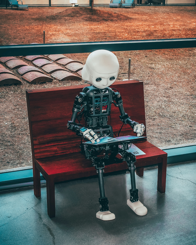 Kleiner Roboter sitzt auf einer Holzbank und liest in einem Heft