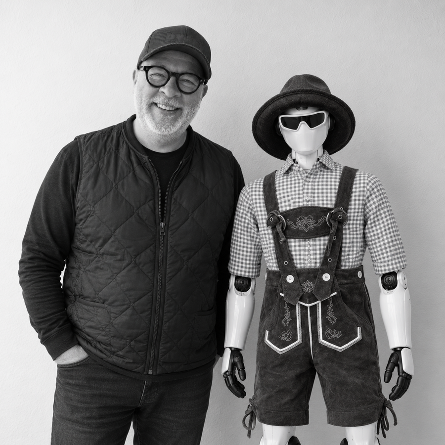 Mann mit Brille und Kappe steht neben einem Roboter in Lederhose und Trachtenhemd