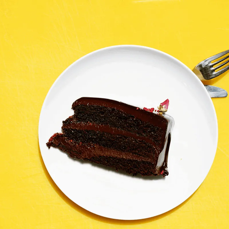 Glänzendes dreischichtiges Schokoladenkuchenstück auf weißem Teller vor gelbem Hintergrund