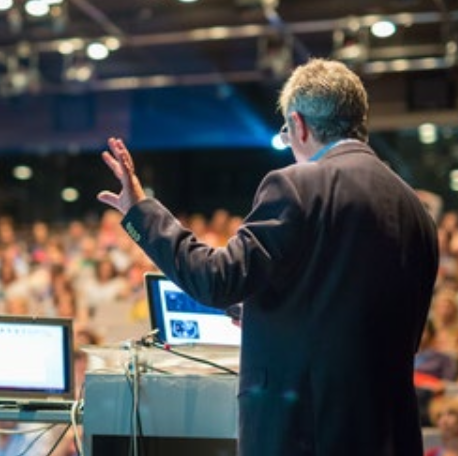 Keynote-Redner auf Konferenzbühne vor aufmerksamem Publikum, Laptops und Bühnentechnik sichtbar
