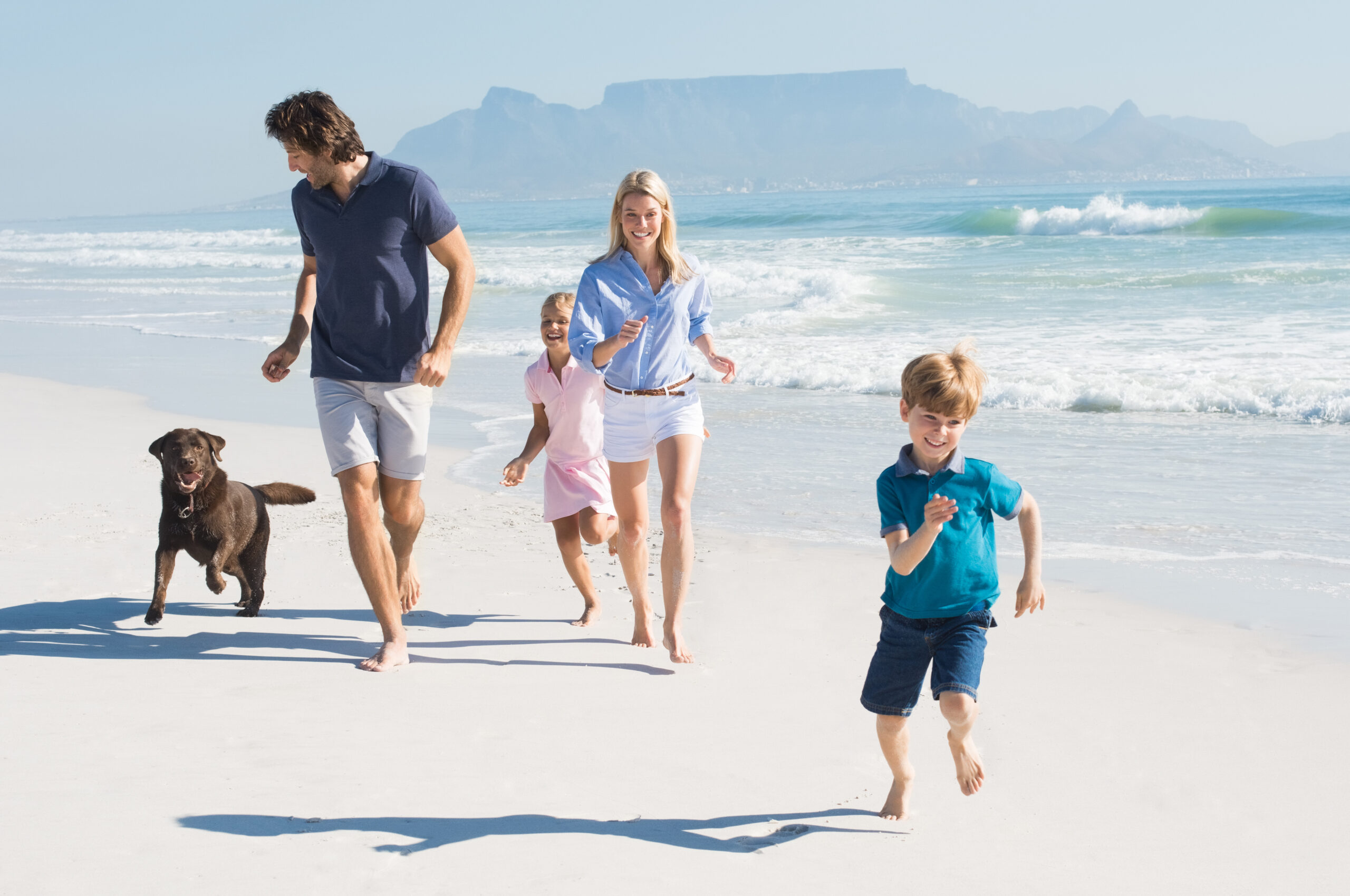 Familie mit Hund und Kindern rennt barfuß am sonnigen Sandstrand am Meer