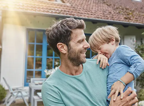 Vater umarmt lachenden Sohn im sonnigen Garten vor blauem Fenster