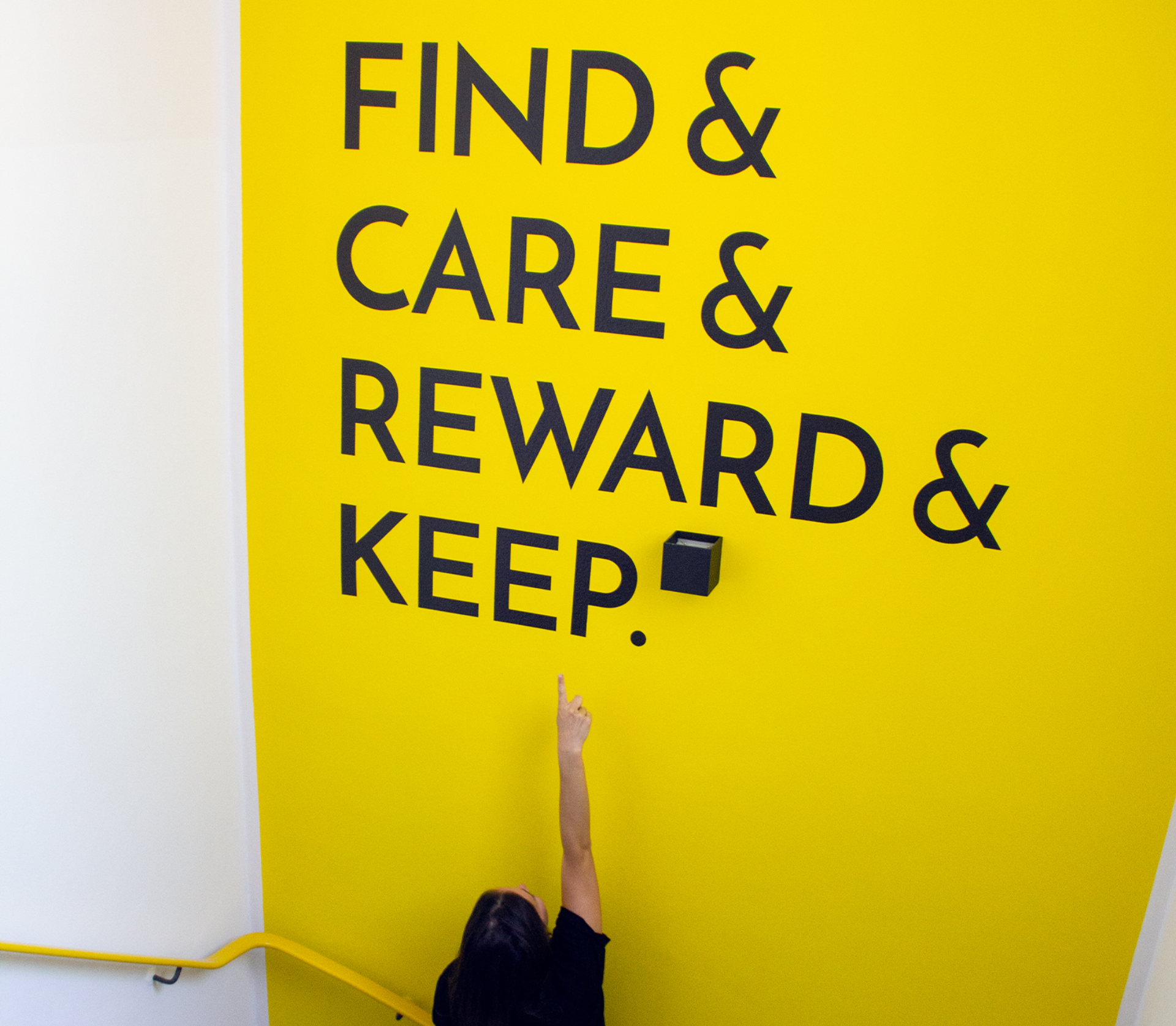 Gelbes Wandbild mit großer schwarzer Typografie FIND & CARE & REWARD & KEEP, Person zeigt.