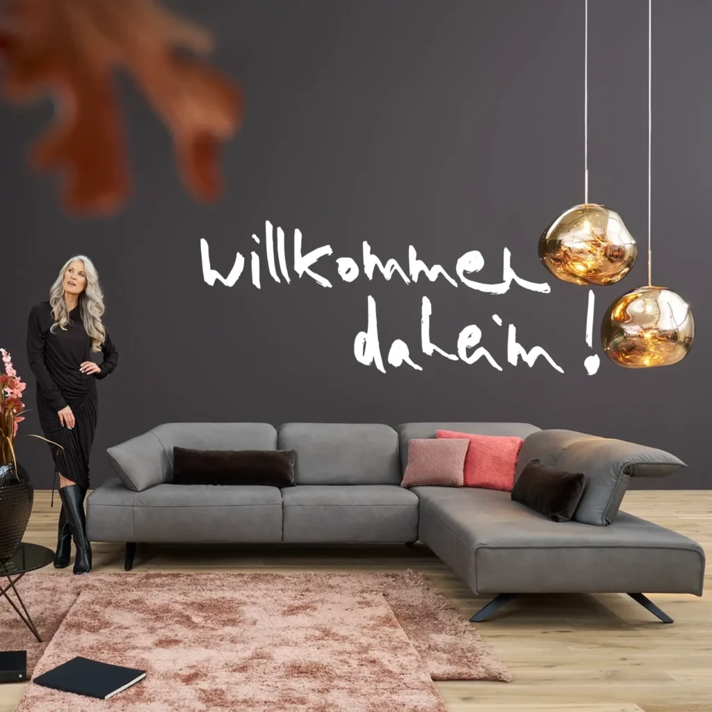 Modernes Wohnzimmer mit grauem Ecksofa, rosa Teppich und Wandtext „Willkommen daheim“.