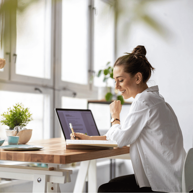 Lächelnde Frau im Homeoffice am Laptop, heller Arbeitsplatz mit Notizbuch, Kaffeetasse und Pflanze