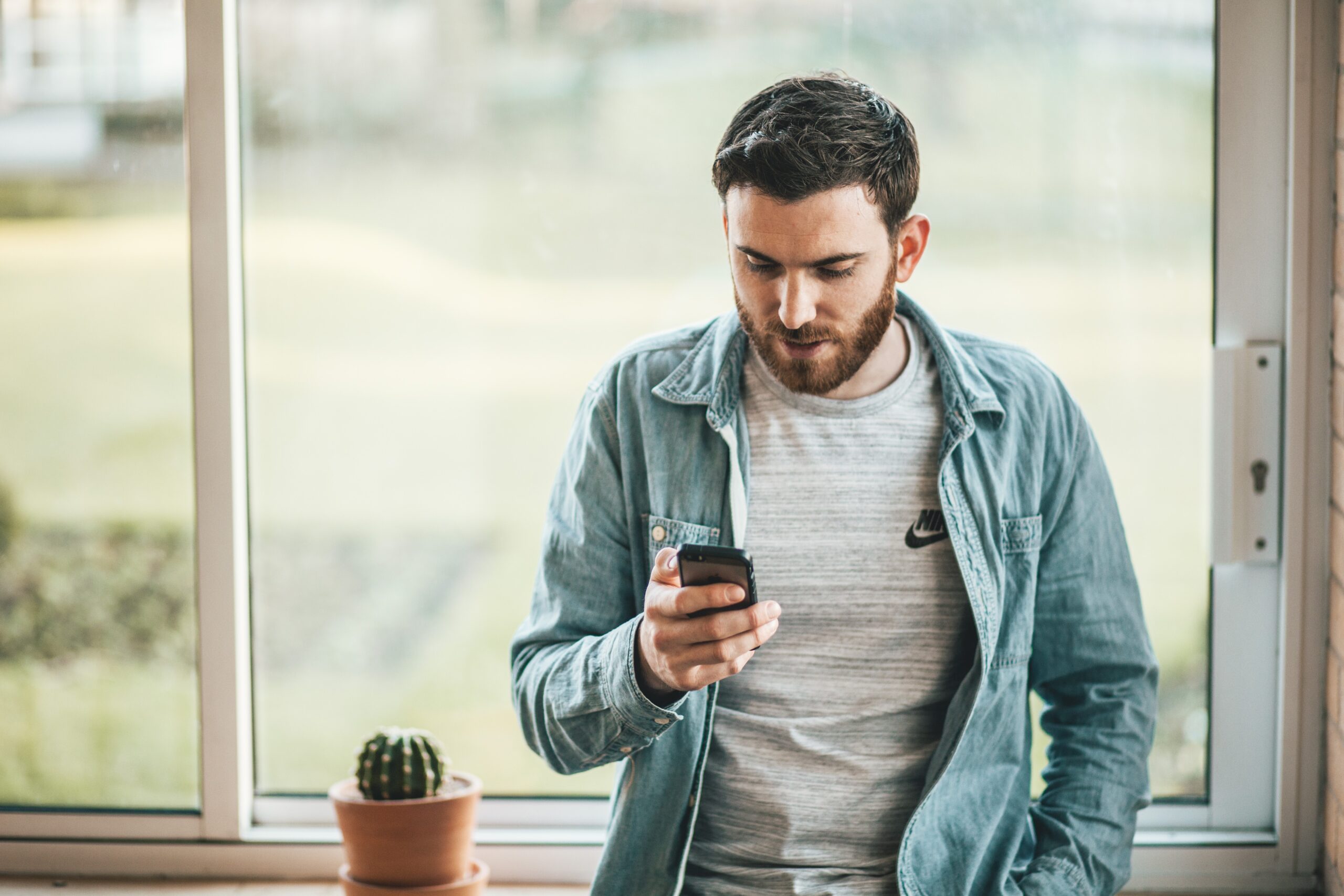 Mann am Fenster mit Smartphone, nachdenklich im natürlichen Licht neben Kaktus