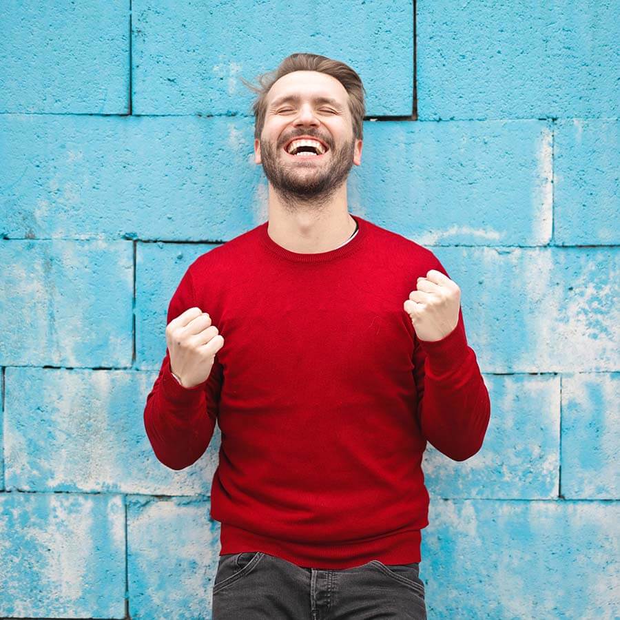 Jubelnder Mann im roten Pullover vor blauer Betonwand, energiegeladenes Triumphgefühl