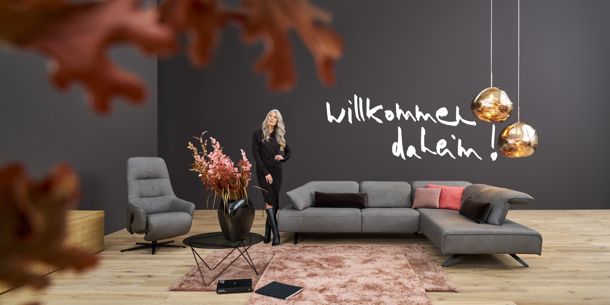 Modernes Wohnzimmer mit grauem Ecksofa, rosafarbenem Teppich und Willkommen daheim-Wandschrift