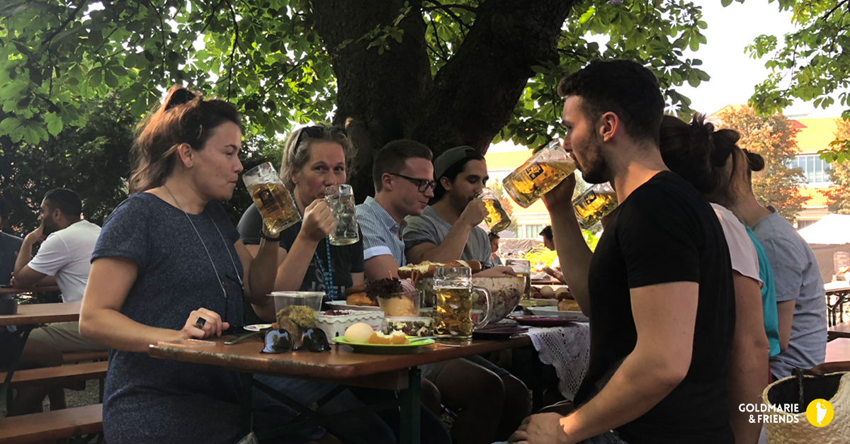 Menschen sitzen im Biergarten an langen Tischen, essen und trinken unter Bäumen