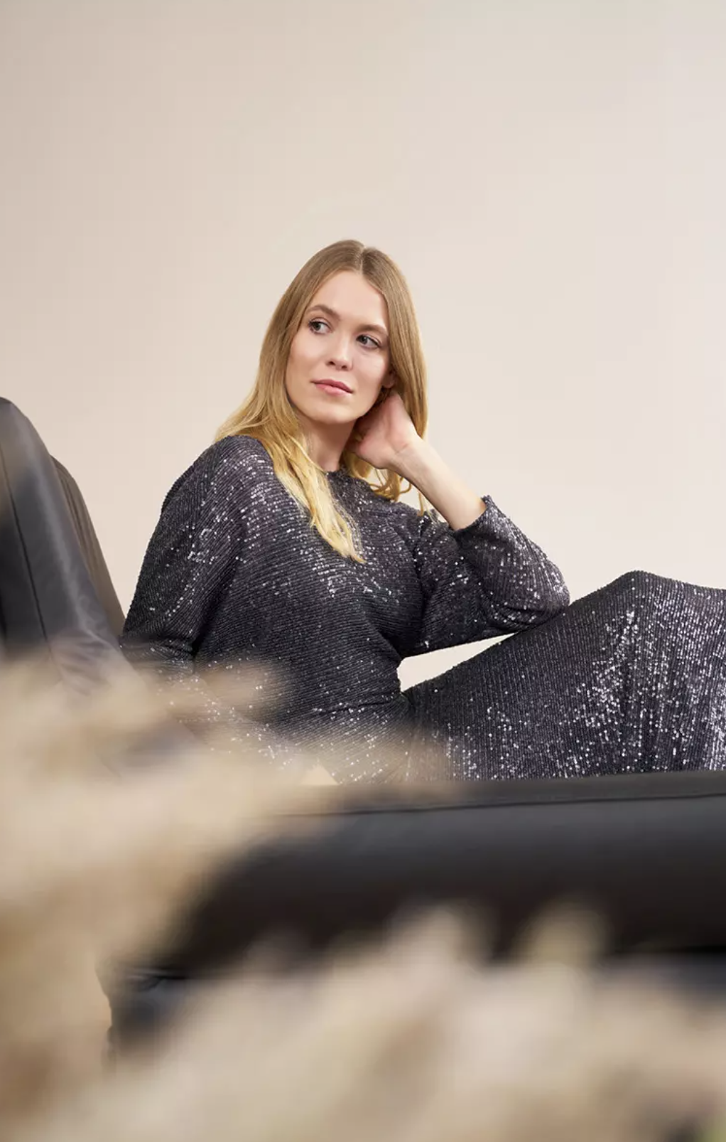 Blonde Frau im glitzernden Abendkleid entspannt auf schwarzem Ledersofa, intimes Porträt