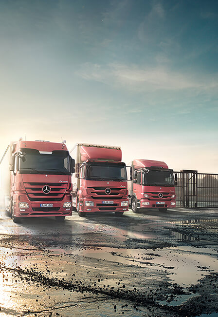 Drei rote Mercedes-Lkw in Flottenformation auf nassem Parkplatz mit Spiegelungen