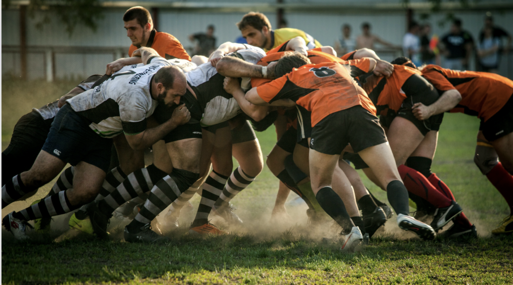 Rugby-Scrum: intensiver Kampf um Ballbesitz, orange gegen weiß, schlammige Trikots, gedrängte Körper