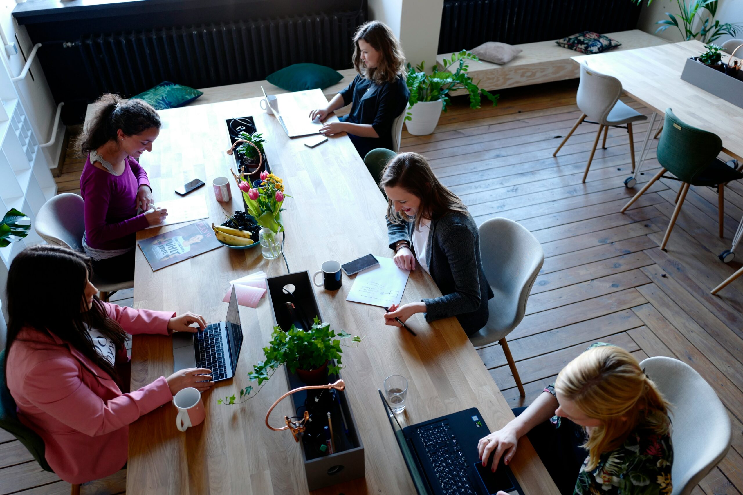 Moderner Coworking-Tisch für Teamarbeit: langer Holztisch mit fünf Arbeitsplätzen, Laptops, Pflanzen