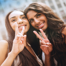 Nahaufnahme: zwei Freundinnen lächelnd im Selfie mit Peace-Zeichen, sonnige Outdoor-Stimmung