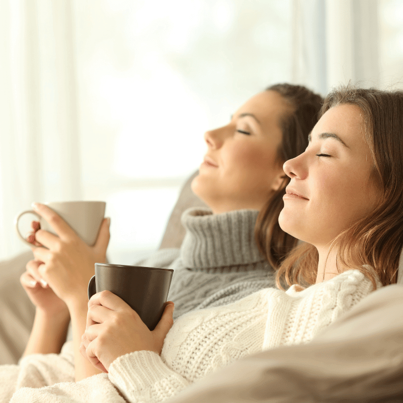 Zwei Frauen auf Sofa mit Tassen, gemütliche sonnige Entspannung in Pullovern