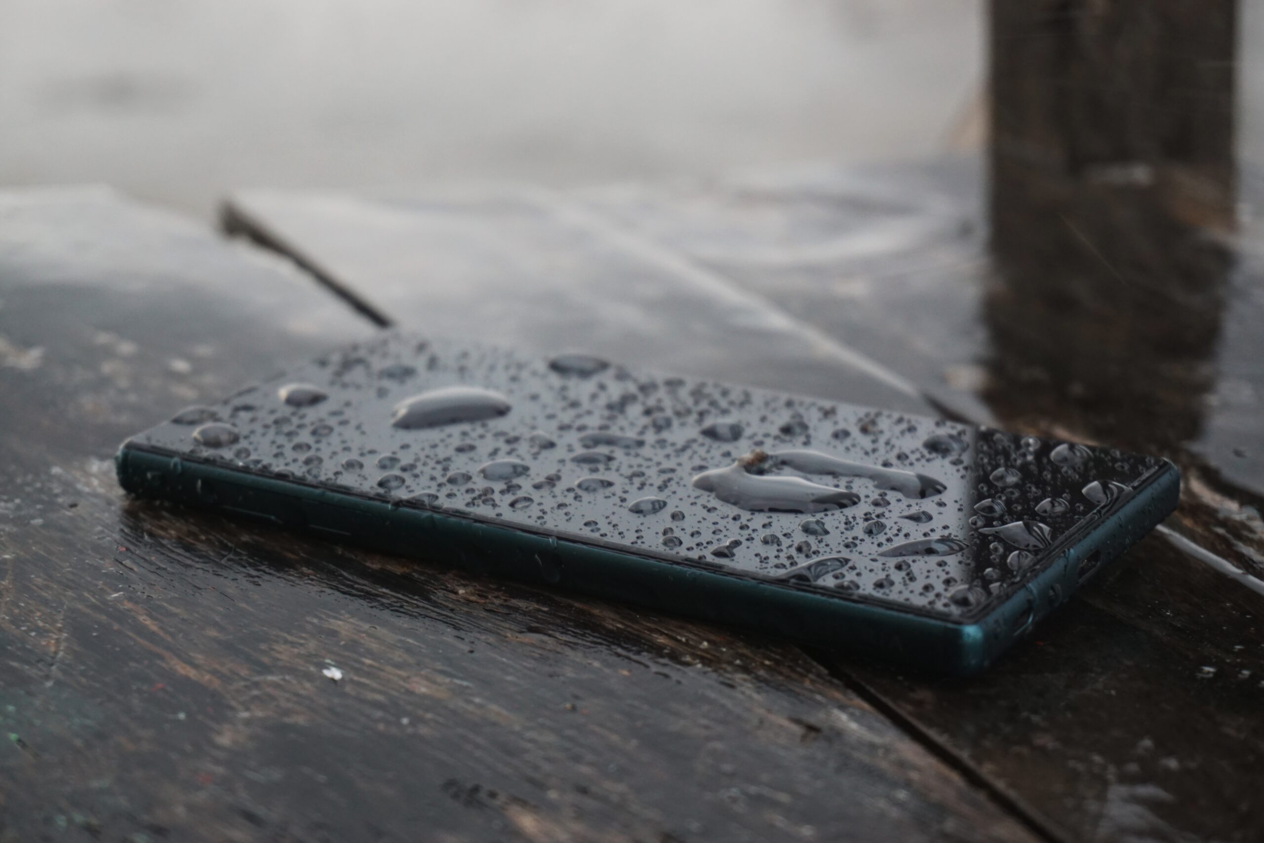Nasses Smartphone mit Regentropfen auf Holz, zwei sichtbare Kameralinsen