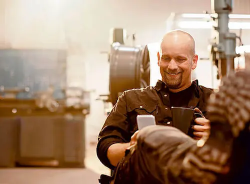 Werkstattpause: lächelnder Arbeiter mit Handy und Thermobecher